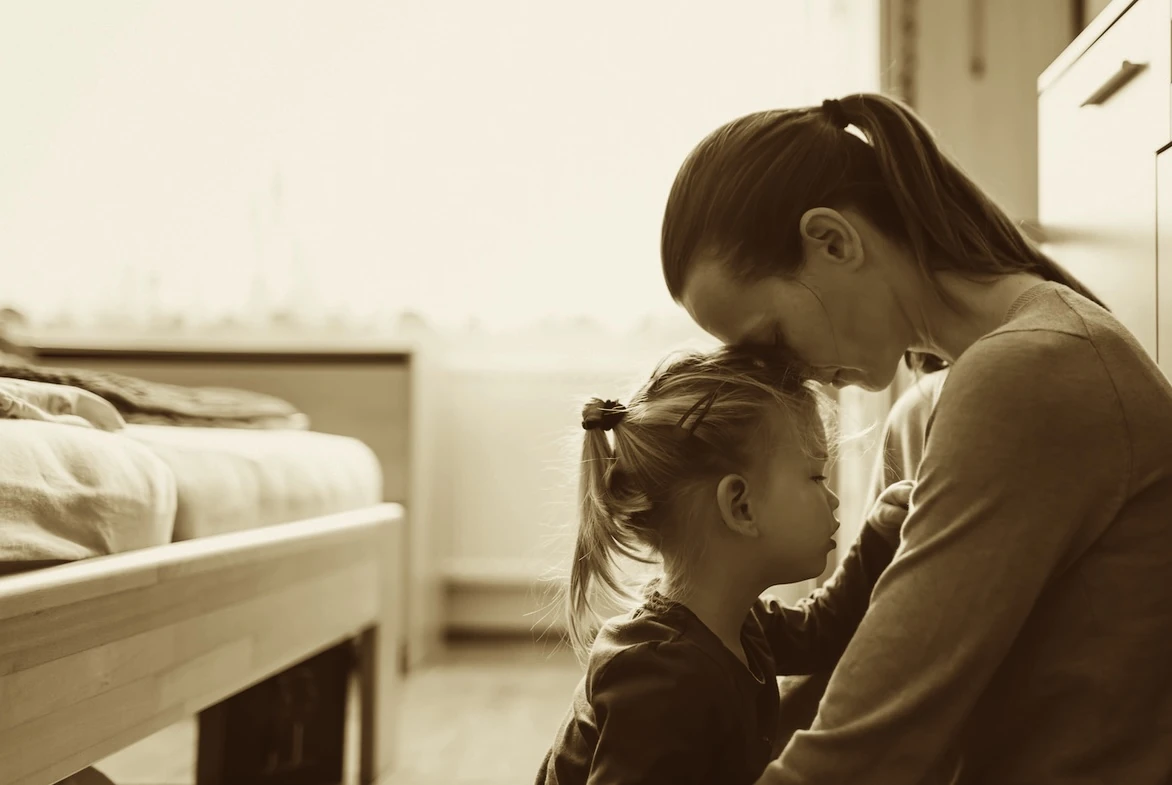A mother resting her forehead against her child’s head as they sit together beside a bed